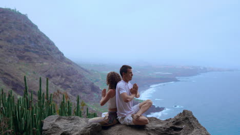Atop-a-mountain,-a-man-and-woman-sit-back-to-back-on-a-rock,-practicing-meditation-and-yoga-while-the-ocean-offers-a-serene-background